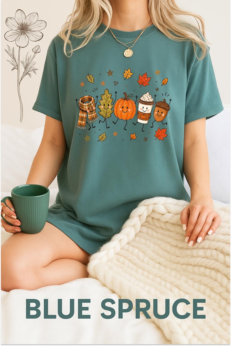 May include: A blue-green short-sleeve t-shirt featuring a graphic print of a smiling scarf, leaf, pumpkin, coffee cup, and acorn dancing together, surrounded by small autumn leaves and stars. The shirt has a crew neckline and a relaxed fit.