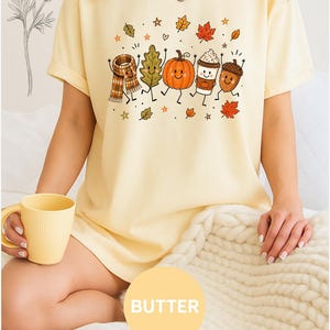May include: A pale yellow t-shirt featuring a graphic print of five anthropomorphic autumn characters including a plaid scarf, a leaf, a pumpkin, a coffee cup, and an acorn, surrounded by small stars and leaves, with the word BUTTER in a circle.