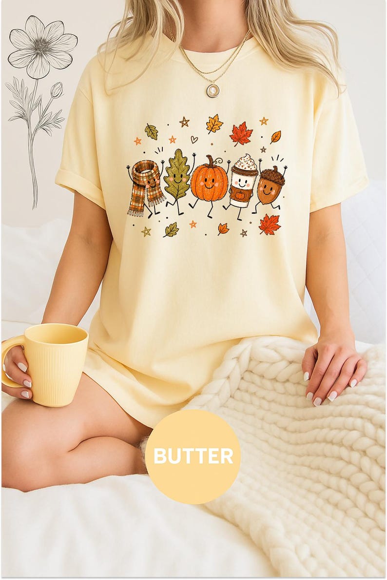 May include: A pale yellow t-shirt featuring a graphic print of five anthropomorphic autumn characters including a plaid scarf, a leaf, a pumpkin, a coffee cup, and an acorn, surrounded by small stars and leaves, with the word BUTTER in a circle.