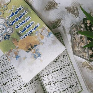 Puede incluir: Un libro abierto con escritura árabe y una cubierta verde y dorada con texto árabe. Un segundo libro con una cubierta verde y texto árabe está encima. Una pequeña planta en un recipiente de vidrio está en el fondo.