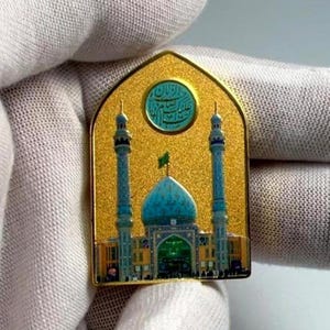 May include: Gold-colored pin featuring a mosque with a blue dome and two minarets. The pin has a textured background and a small green flag. A circular emblem with Arabic script is at the top.