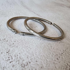 May include: Two silver-colored metal shower curtain rings. Each ring is a semi-circle with a pointed end. The rings are lying on a textured, off-white surface. The rings are slightly overlapping.
