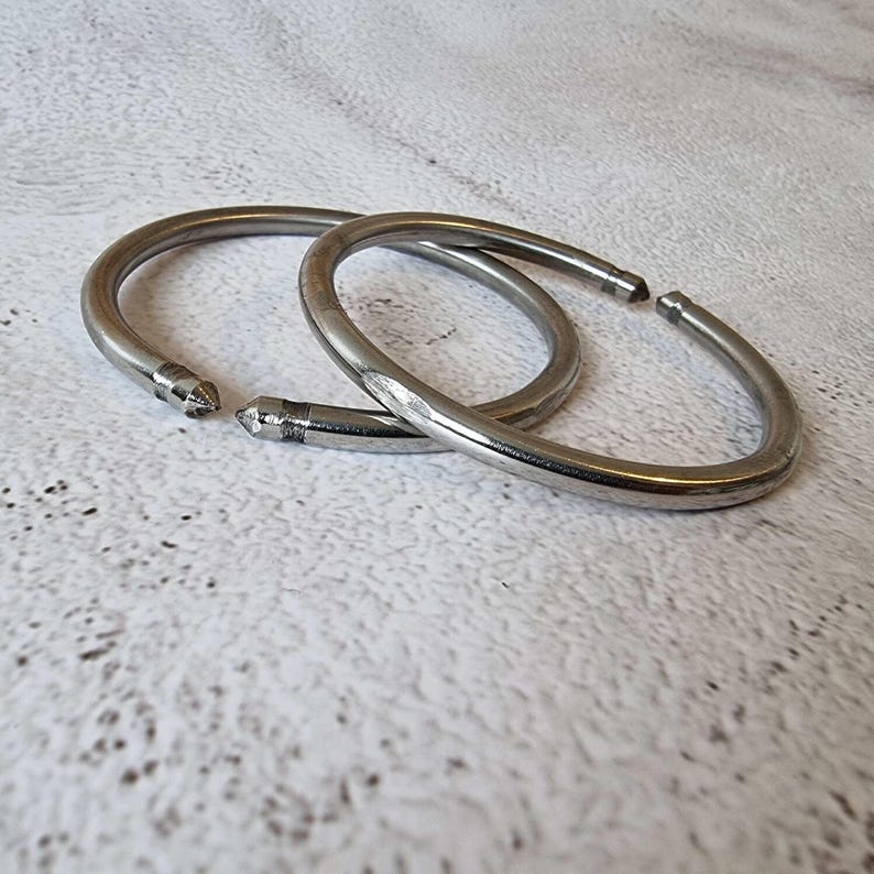 May include: Two silver-colored metal shower curtain rings. Each ring is a semi-circle with a pointed end. The rings are lying on a textured, off-white surface. The rings are slightly overlapping.