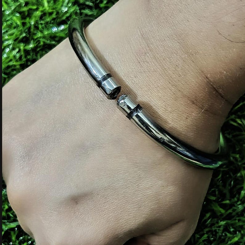 May include: A silver-colored, hinged bangle bracelet. The bracelet is made of a thick, rounded metal and features a clasp that opens and closes. The bracelet is shown on a person's wrist against a green background.