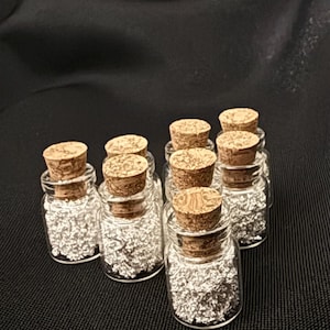 May include: A collection of small, clear glass bottles with cork stoppers. Each bottle is filled with silver-colored, glitter-like material. The bottles are arranged on a dark surface.