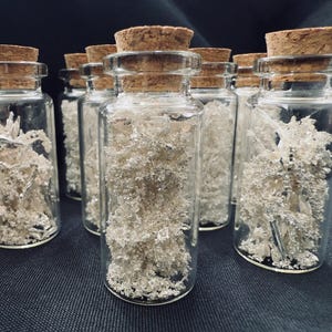 May include: A collection of small, clear glass bottles with cork stoppers. Each bottle is filled with a cluster of silver-colored, crystalline material. The bottles are arranged on a dark surface, creating a visually appealing display.