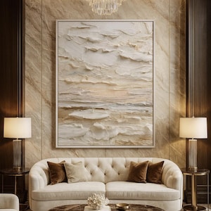 Puede incluir: Una obra de arte abstracta texturizada enmarcada, en tonos neutros, cuelga sobre un sofá crema capitoné con cojines marrones. Dos lámparas a juego flanquean el sofá, y una mesa de centro redonda de mármol se encuentra delante.