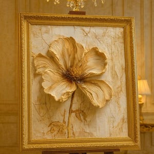 May include: A framed, textured artwork featuring a large, three-dimensional flower in shades of gold and cream. The artwork is displayed on a wooden easel, with a gold chandelier in the background. The frame is gold.