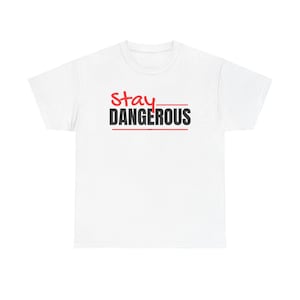 Puede incluir: Camiseta blanca con la frase "Stay DANGEROUS" impresa en negro y rojo. La palabra "Stay" está en un estilo manuscrito, mientras que "DANGEROUS" está en negrita, con una fuente sans-serif. Una línea roja subraya la palabra "DANGEROUS".