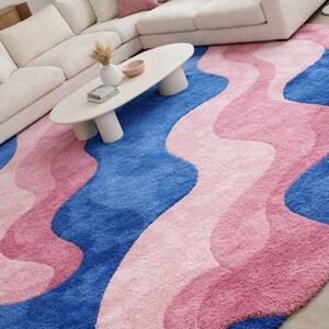May include: A large, plush area rug with a wavy design in shades of blue and pink. The rug features a vibrant blue base with flowing, organic shapes in varying shades of pink. The rug is placed in a living room setting, with a white sofa and coffee table.