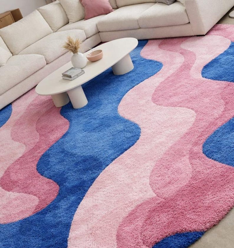 May include: A large, plush area rug with a wavy design in shades of blue and pink. The rug features a vibrant blue base with flowing, organic shapes in varying shades of pink. The rug is placed in a living room setting, with a white sofa and coffee table.