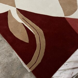 Pu&ograve; includere: Un tappeto moderno con un design astratto. Il tappeto presenta uno sfondo bordeaux scuro con forme geometriche color crema, beige e bordeaux chiaro. Il design del tappeto &egrave; contemporaneo e si adatterebbe a uno spazio abitativo moderno.