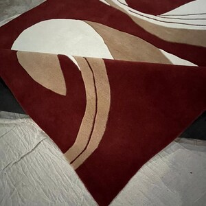 Pu&ograve; includere: Un tappeto bordeaux con un design astratto. Il tappeto presenta grandi forme curve in bianco sporco, beige e bordeaux. Il tappeto &egrave; piegato, rivelando il suo rovescio.