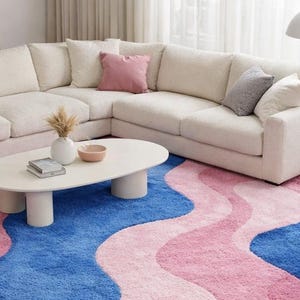 May include: A modern living room setup featuring a large, off-white sectional sofa with several throw pillows. A white oval coffee table sits on a vibrant rug with blue and pink wavy patterns. A floor lamp provides soft lighting.