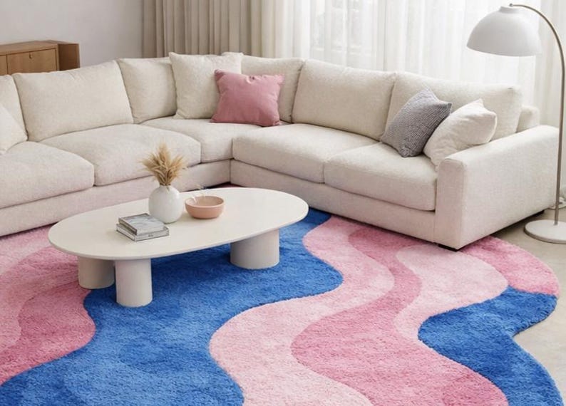 May include: A modern living room setup featuring a large, off-white sectional sofa with several throw pillows. A white oval coffee table sits on a vibrant rug with blue and pink wavy patterns. A floor lamp provides soft lighting.