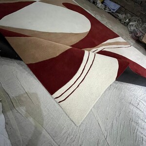 Pu&ograve; includere: Un grande tappeto arrotolato con un design moderno. Il tappeto presenta forme geometriche bordeaux, crema e beige. Il tappeto &egrave; realizzato in un materiale morbido. Il design &egrave; astratto e adatto per un soggiorno o una camera da letto.