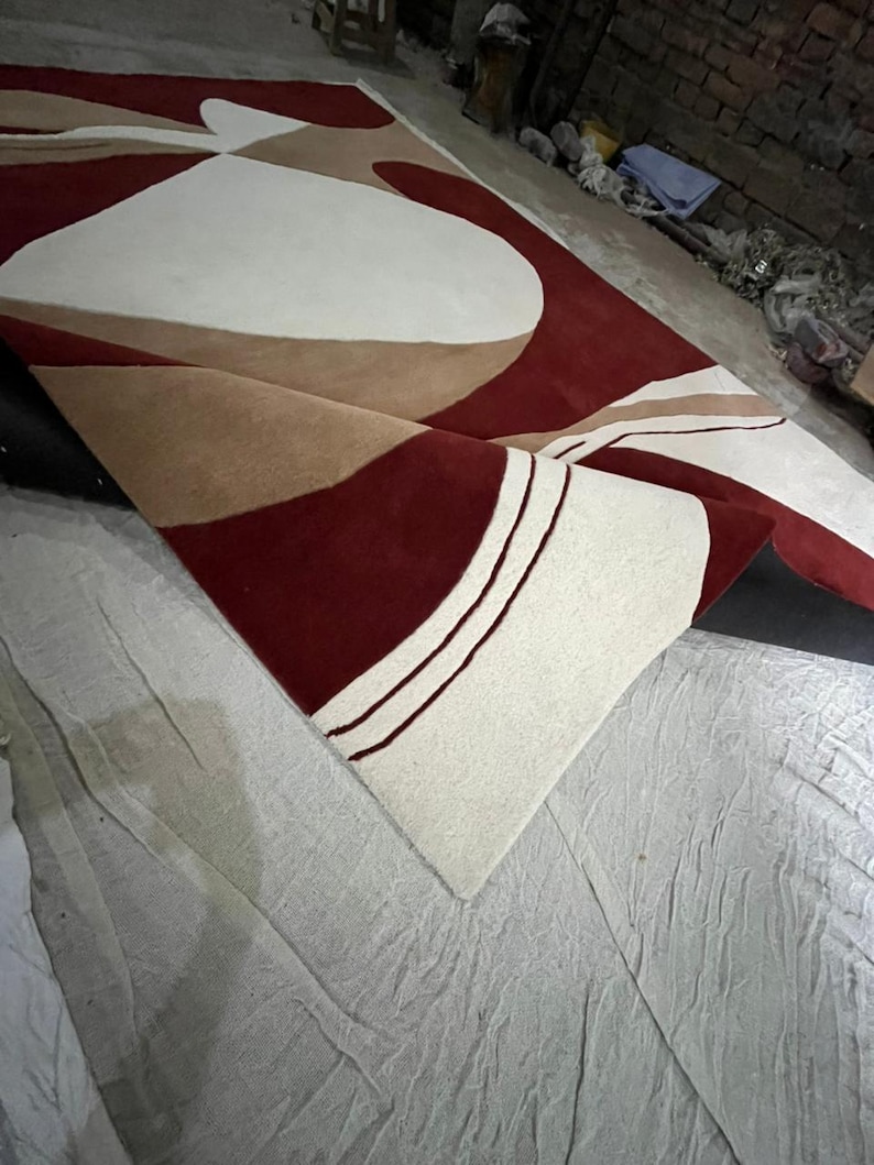 Pu&ograve; includere: Un grande tappeto arrotolato con un design moderno. Il tappeto presenta forme geometriche bordeaux, crema e beige. Il tappeto &egrave; realizzato in un materiale morbido. Il design &egrave; astratto e adatto per un soggiorno o una camera da letto.