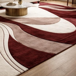 Pu&ograve; includere: Un grande tappeto con un design astratto nei colori bordeaux, beige e marrone chiaro. Il motivo presenta linee e forme curve, creando un'estetica moderna. Il tappeto &egrave; posato su un pavimento in legno.