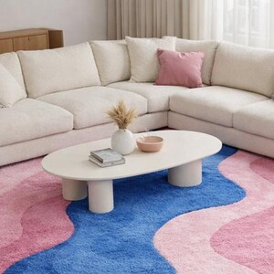 May include: A cream-colored sectional sofa with several throw pillows sits on a blue and pink wavy rug. A white oval coffee table with cylindrical legs holds a vase, bowl, and books. A wooden cabinet and a floor lamp are also visible.