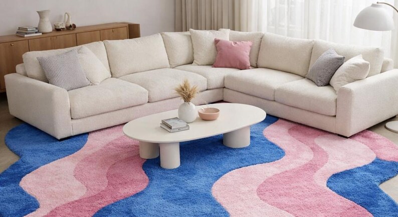 May include: A cream-colored sectional sofa with several throw pillows sits on a blue and pink wavy rug. A white oval coffee table with cylindrical legs holds a vase, bowl, and books. A wooden cabinet and a floor lamp are also visible.