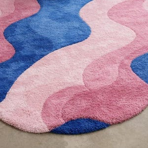 May include: A plush, wavy rug in shades of blue, pink, and rose. The rug has a soft, textured surface and a unique, organic shape. The design features flowing, layered color blocks, adding a modern touch to any space.