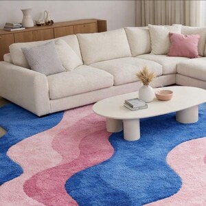 May include: A modern living room scene featuring a large, off-white sectional sofa, a white oval coffee table, and a vibrant rug. The rug has a wavy design in shades of blue and pink. Decorative pillows and vases add pops of color.