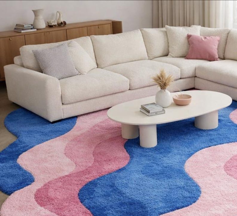 May include: A modern living room scene featuring a large, off-white sectional sofa, a white oval coffee table, and a vibrant rug. The rug has a wavy design in shades of blue and pink. Decorative pillows and vases add pops of color.