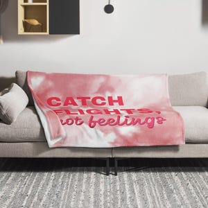 Peut inclure: Une couverture rose et blanche avec le texte "CATCH FLIGHTS, not feelings" en lettres rouges et roses. La couverture est drapée sur un canapé gris dans un salon.