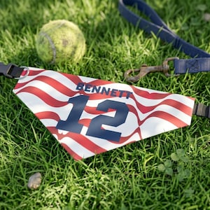 Op de afbeelding: Een gepersonaliseerde hondenbandana met een patriottisch ontwerp, met rode en witte strepen, het nummer 12 in marineblauw en de naam "BENNETT". Een tennisbal en een blauwe riem met metalen sluiting zijn ook zichtbaar.