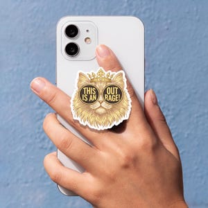 May include: A pop-up phone grip featuring a golden cat wearing sunglasses and a crown. The text on the grip reads "THIS IS AN OUT RAGE!". The cat has a fluffy appearance and is set against a blue background.