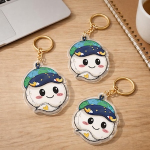 May include: Three clear acrylic keychains featuring a cartoon character with a white body, a blue and green hat, and a gold keychain ring. The character has a smiling face with black eyes and a small yellow heart.