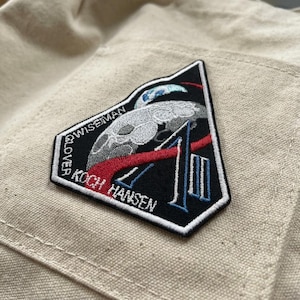 May include: Embroidered patch featuring a stylized moon and a rocket, with the number "1" in blue. The patch includes the names "Glover", "Wiseman", "Koch", and "Hansen". The patch is black, white, red, and blue.