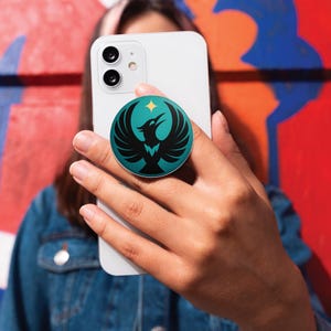 May include: A teal and black phone grip featuring a stylized bird design with outstretched wings and a star. The phone accessory is attached to a white smartphone, held against a colorful background.