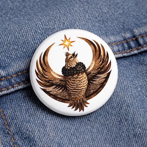 May include: A white button featuring a detailed illustration of a brown owl with outstretched wings and a star above its head. The owl has a black and brown feathered collar. The button is set against a blue denim background.