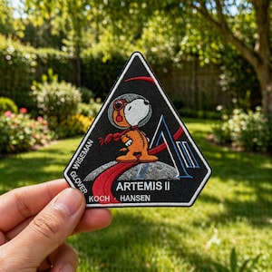 May include: A black, triangular patch featuring Snoopy in an orange spacesuit, standing on a moon with the text "ARTEMIS II". The patch includes the names "WISEMAN", "GLOVER", "KOCH", and "HANSEN". A red and blue design is also present.