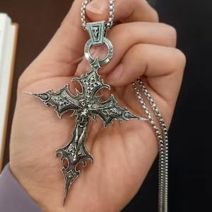 Może przedstawiać: Srebrny naszyjnik z wisiorkiem z ozdobnym krzyżem. Krzyż ma postać krucyfiksu i skomplikowane wzory. Naszyjnik jest trzymany w dłoni, prezentując łańcuszek i gotycki styl wisiorka.