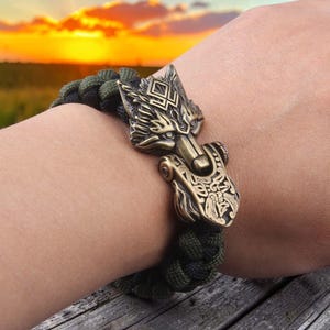 Puede incluir: Una pulsera trenzada verde y negra con un cierre de cabeza de lobo color bronce. La cabeza de lobo presenta detalles intrincados y un diseño decorativo. La pulsera se muestra en una muñeca, con una puesta de sol al fondo.