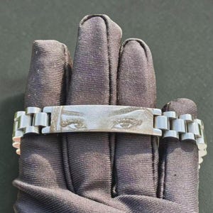 May include: A silver bracelet with a rectangular central plate engraved with an image of eyes. The bracelet has a linked band design. The bracelet is displayed on a dark fabric surface.