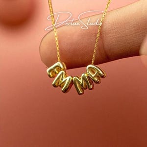 Könnte beinhalten: Eine goldfarbene Halskette mit den Buchstaben "EMMA", die einen Namen bilden. Die Buchstaben sind einzeln, abgerundet und durch eine zarte Kette verbunden. Der Hintergrund ist einfarbig warmrosa.