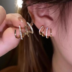 May include: Gold-toned earrings with a claw-like design, adorned with small, sparkling crystals. One earring is being held up, while another is worn on the ear. The earrings have a curved, three-prong design, creating a modern and elegant look.