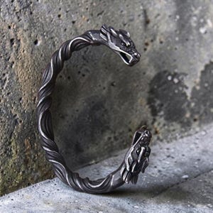 Puede incluir: Una pulsera de puño plateada con un diseño de dragón. La pulsera presenta una banda retorcida y cabezas de dragón en cada extremo. Las cabezas de dragón tienen escamas detalladas y bocas abiertas. La pulsera es de metal y tiene un aspecto envejecido.