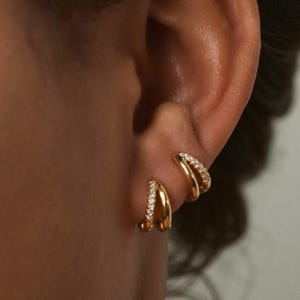 May include: A close-up of two gold-colored hoop earrings, each adorned with small, sparkling gemstones. The earrings are worn on an ear, showcasing their design and the way they sit on the earlobe. The jewelry has a polished finish.