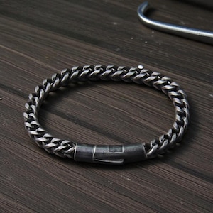 Puede incluir: Una pulsera de cadena de color plateado con un cierre seguro. La pulsera está hecha de eslabones entrelazados y se muestra sobre una superficie de madera oscura. Un accesorio elegante.