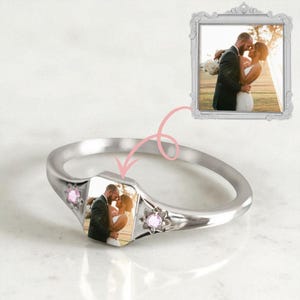 May include: A silver ring with a rectangular photo insert featuring a couple. The ring has two pink gemstones and a decorative frame with a photo of the same couple. The ring is on a white surface.