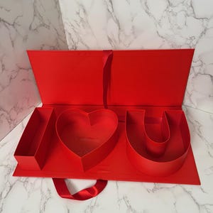 May include: A red gift box with the letters "I", a heart, and "U" arranged inside. The box is open, revealing the shapes, and has a red ribbon handle. The background is a white marble pattern.