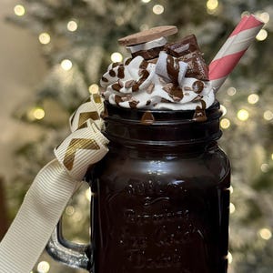 May include: A dark brown mason jar filled with a chocolate drink, topped with whipped cream, chocolate shavings, and pieces of chocolate and a cookie. A pink and white striped straw and a cream ribbon with gold accents complete the look.