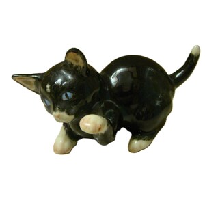 May include: A small ceramic cat figurine with a glossy black coat, white paws, and a white chin. The cat is depicted in a playful, crouching posture with one front paw raised, featuring pointed ears and light blue eyes with a white tail tip.