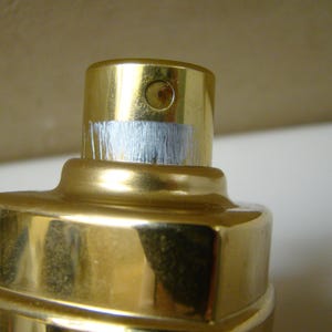 Puede incluir: Primer plano de un frasco de perfume dorado con una banda plateada met&aacute;lica alrededor de la parte superior. El frasco tiene una superficie brillante y reflectante. La parte superior del frasco tiene una peque&ntilde;a abertura circular.