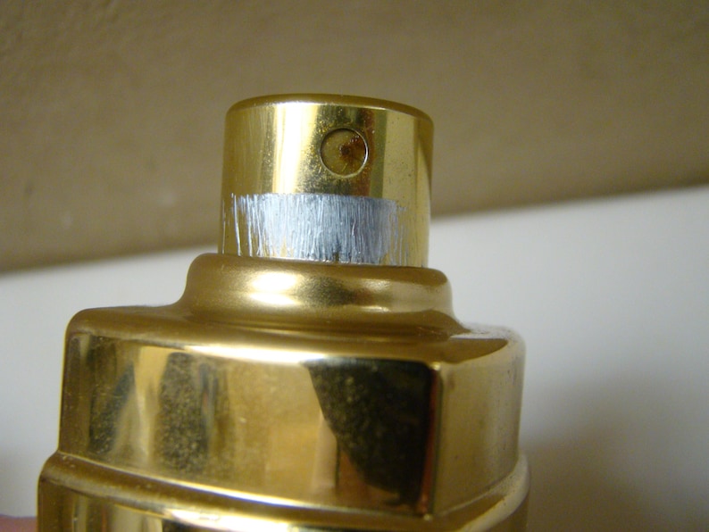 Puede incluir: Primer plano de un frasco de perfume dorado con una banda plateada met&aacute;lica alrededor de la parte superior. El frasco tiene una superficie brillante y reflectante. La parte superior del frasco tiene una peque&ntilde;a abertura circular.