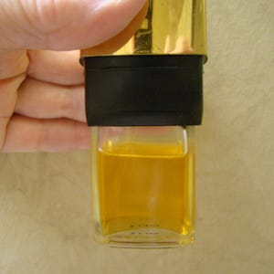 Puede incluir: Una peque&ntilde;a botella de vidrio transparente llena de un l&iacute;quido amarillo dorado. La botella tiene una banda negra y una tapa dorada. La botella est&aacute; sujeta por una mano.